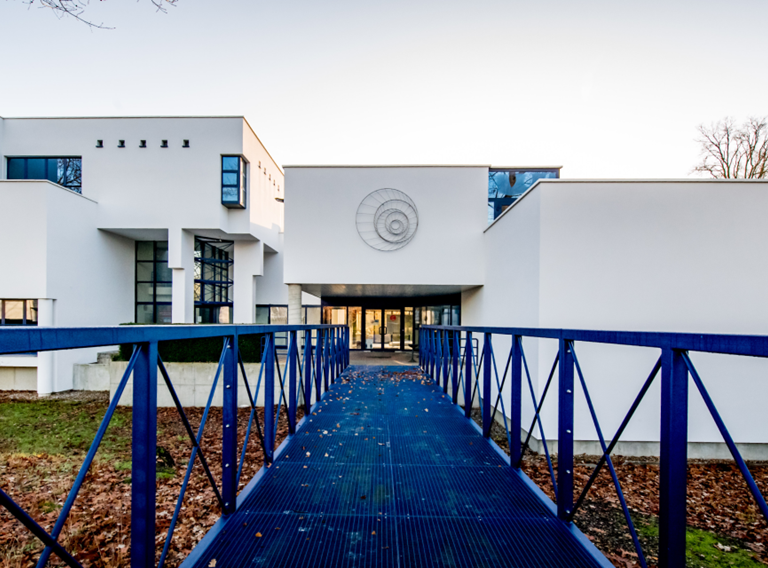 Ons gebouw: een blauwe, metalen loopbrug brengt je naar de ingang van ons modern wit gebouw met spiegelende ramen