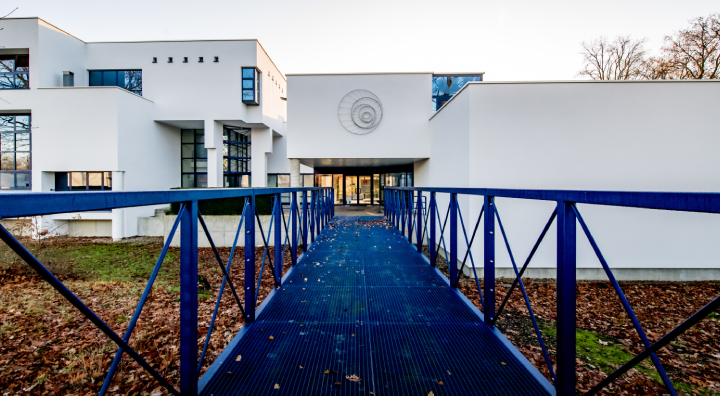 Ons gebouw: een blauwe, metalen loopbrug brengt je naar de ingang van ons modern wit gebouw met spiegelende ramen